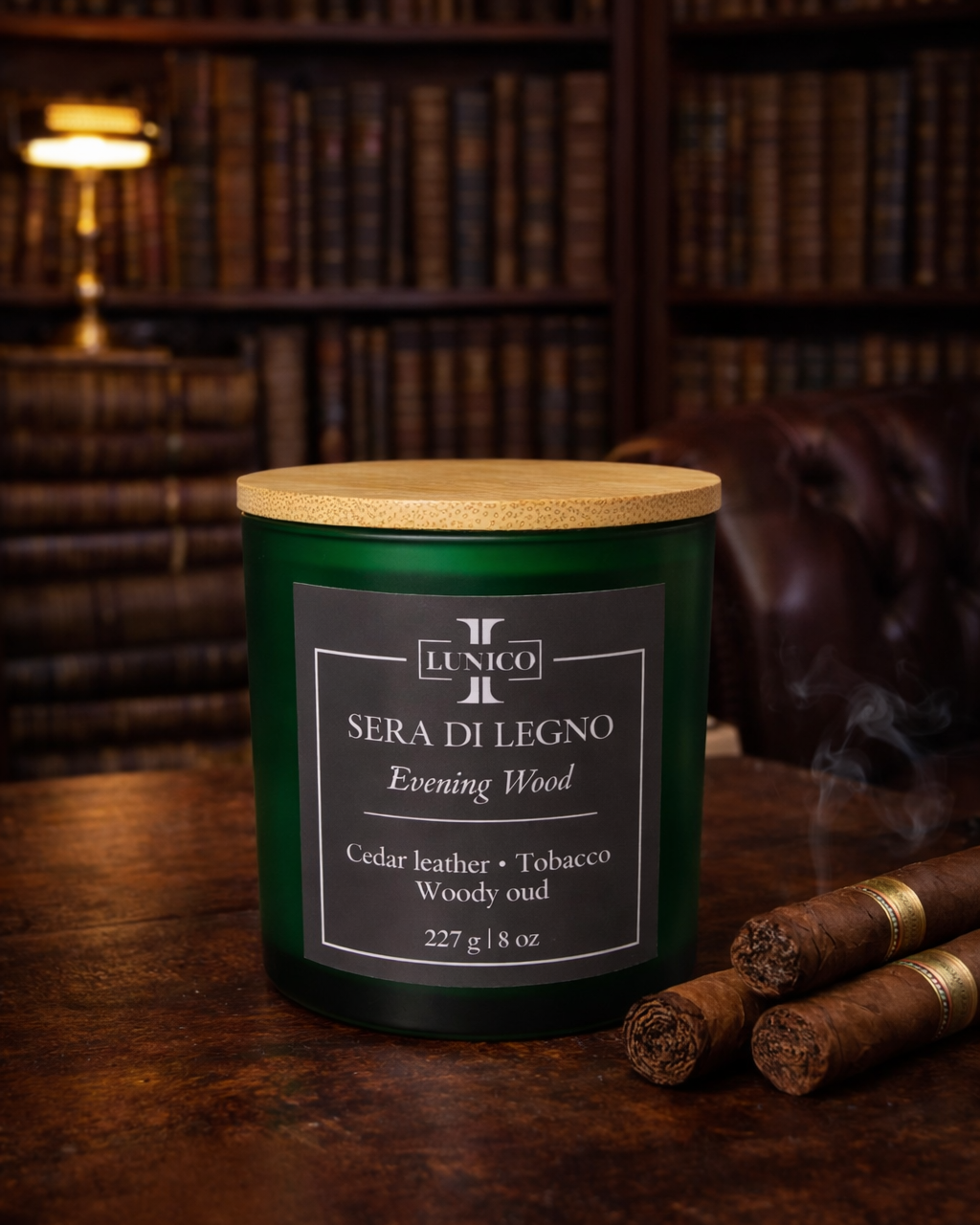 Candle labeled 'Sera di Legno' on a wooden surface with cigars and a bookshelf in the background